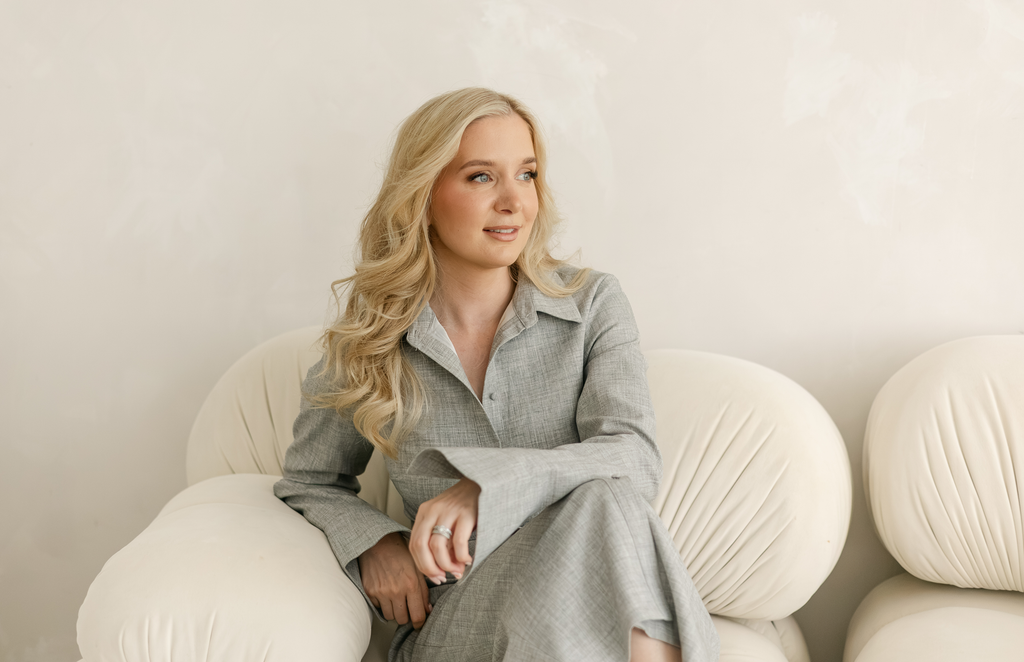 Rachel Harris, sitting on a cream sofa in a grey shirt dress and looking off to the side smiling.