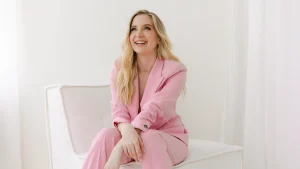 Rachel Harris in pink suit sitting on an off-white sofa in a white room with arms crossed